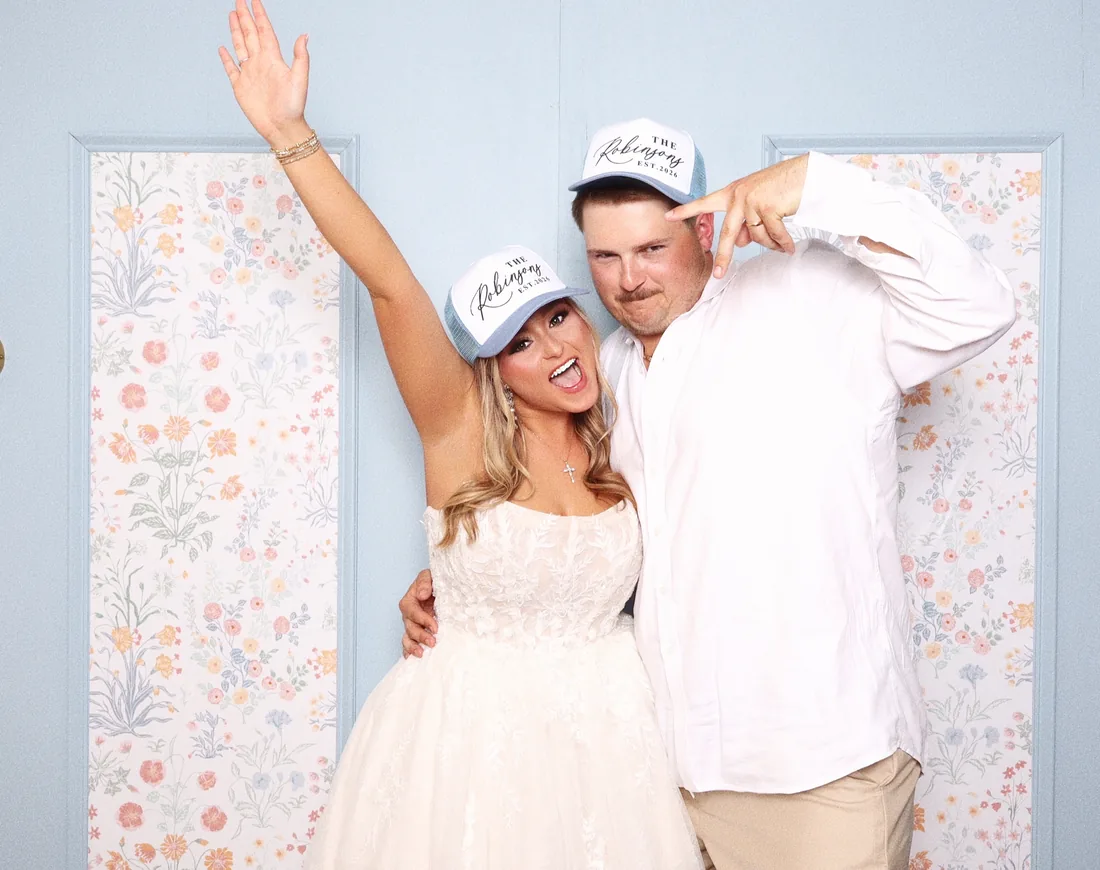 Bride and groom cheering in the booth in front of a pale blue wall papered with flowers.