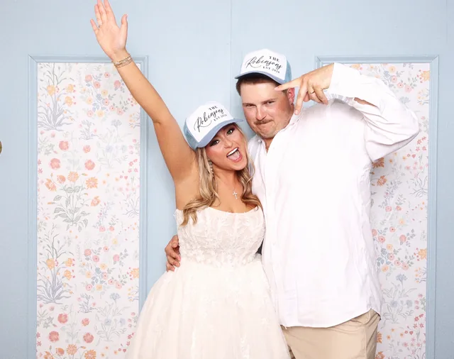 Bride and groom cheering in the booth in front of a pale blue wall papered with flowers.