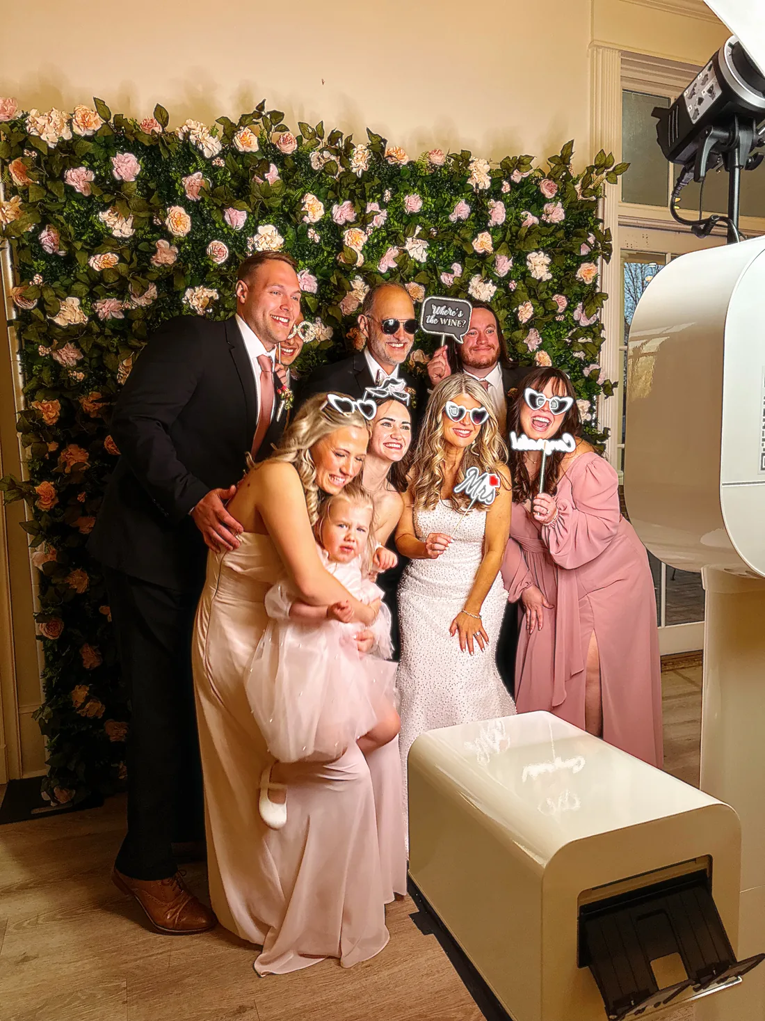 The open air booth and its lighting set up beside a flower wall, with a wedding party posing in front of it.