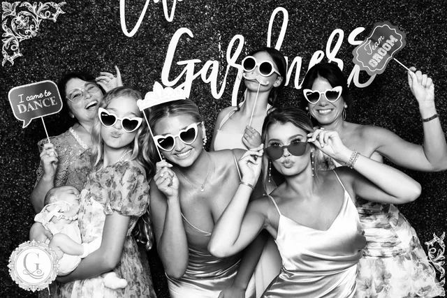 Wedding guests crowded in with props against a dark sequin wall, the couple's monogram in the corner of the frame.
